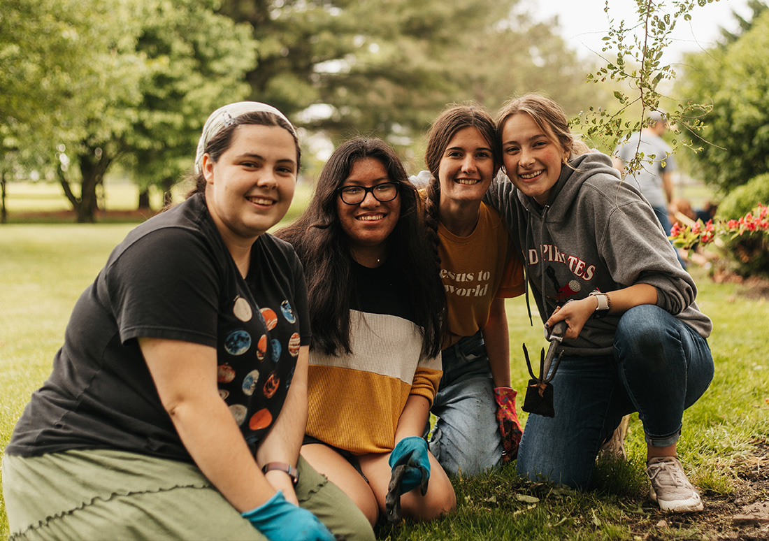 Four women with garden tools and gloves kneel and smile while cleaning up flowerbeds on the RBC campus during Service Day