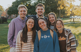 A group of students smile and pose in a group with trees and grass in the background
