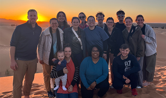 A group of RBC students and a RBC faculty member stand posing in front of a sunset over the desert